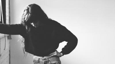 Girl looking down with arm reaching out to nearby window and other hand on hip
